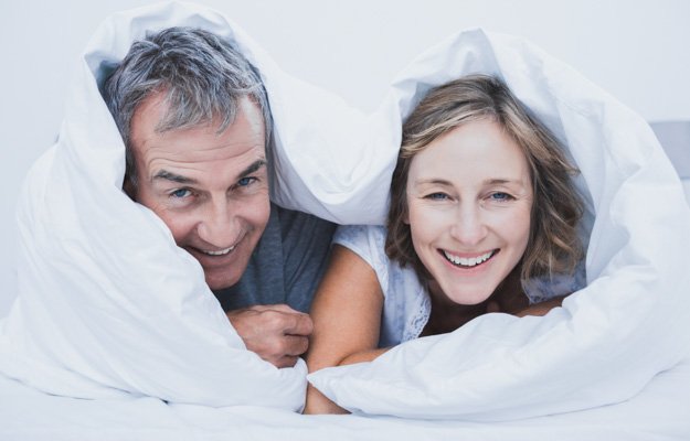 Happy middle aged couple under the duvet smiling at camera in bedroom at home | Low Testosterone Symptoms in Men