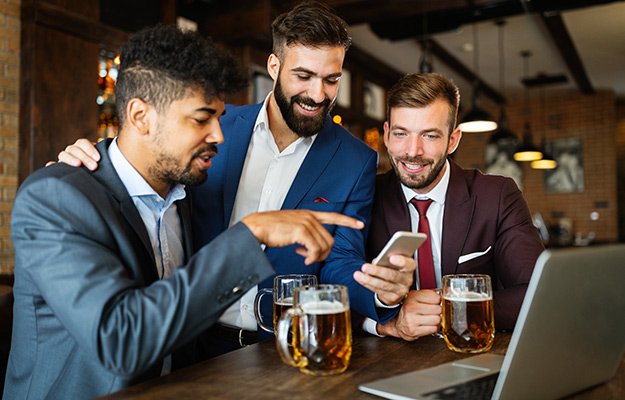 A group of business men drinking beer, looking at phone and laptop | Why Alcohol Hinders Weight Loss